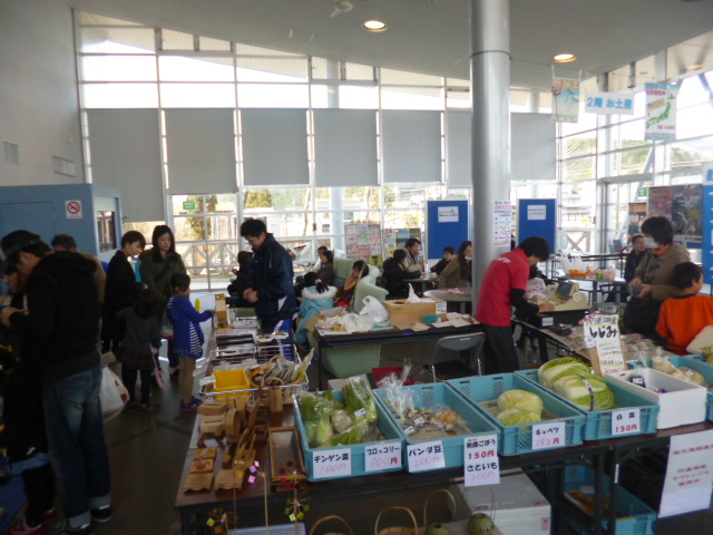 道の駅の施設内にて、採れたての野菜が数多く並ぶ市場とそれを買いに来た人々の写真
