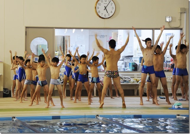 プールの前で水泳のコーチが水着姿の子供たちと一緒に手を上に上げている様子を撮影した写真