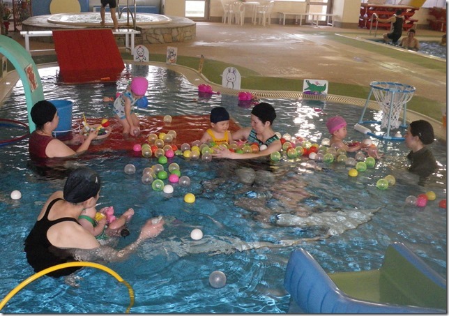 プールの上に浮かぶボールとそのボールを入れる小さな網かごの周りで、複数人のインストラクターと子供が戯れている様子の写真