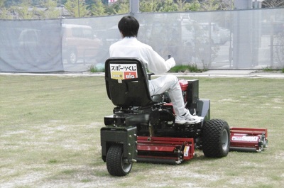 前にタイヤが二つ、後ろにタイヤが一つ付いた赤い車体の芝刈り機に、作業員が乗って操縦し、グラウンドの芝生を整えている様子を斜め後ろから撮影した写真