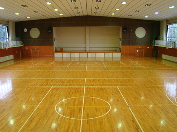 照明が灯った白い天井にバスケットコートが2面設置された玉湯体育館の内観写真、奥にはステージが見える。