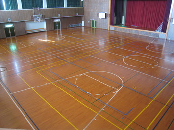 奥に赤いカーテンのある舞台があり、バスケットコートが2面ある体育室を斜め上から撮影した写真