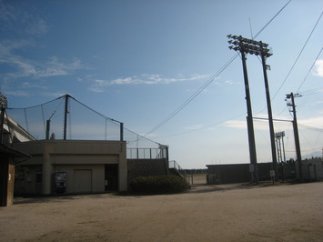晴れた空を背景に、ナイター用の照明がそびえ立ち、周囲を防球ネットで囲まれた八束総合運動場を撮影した写真