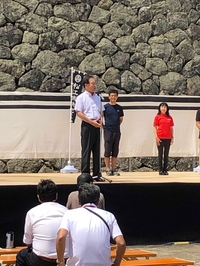 星野副市長が松江城の屋外のステージの上に立ち挨拶をしていて、観客が座って聞いている写真