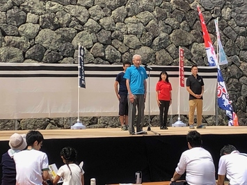 長岡実行委員長が松江城の屋外のステージの上に立ち挨拶をしていて、観客が座って聞いている写真