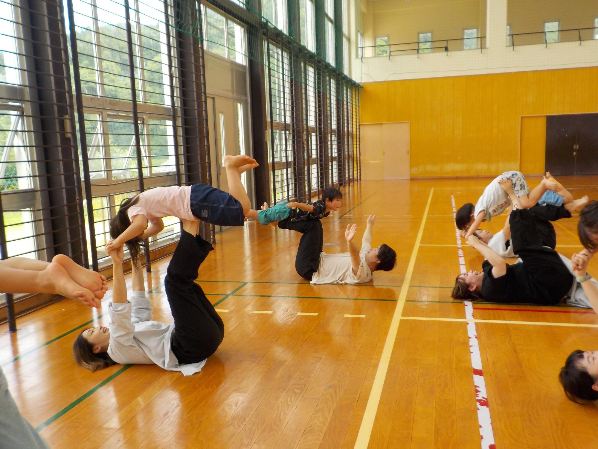 体育館で3組の親子が遊んでいる。親は床に背をつけ、両手両足を上に伸ばしている。こどもは親の手や足の上にうつぶせに乗り、手足を伸ばしている。