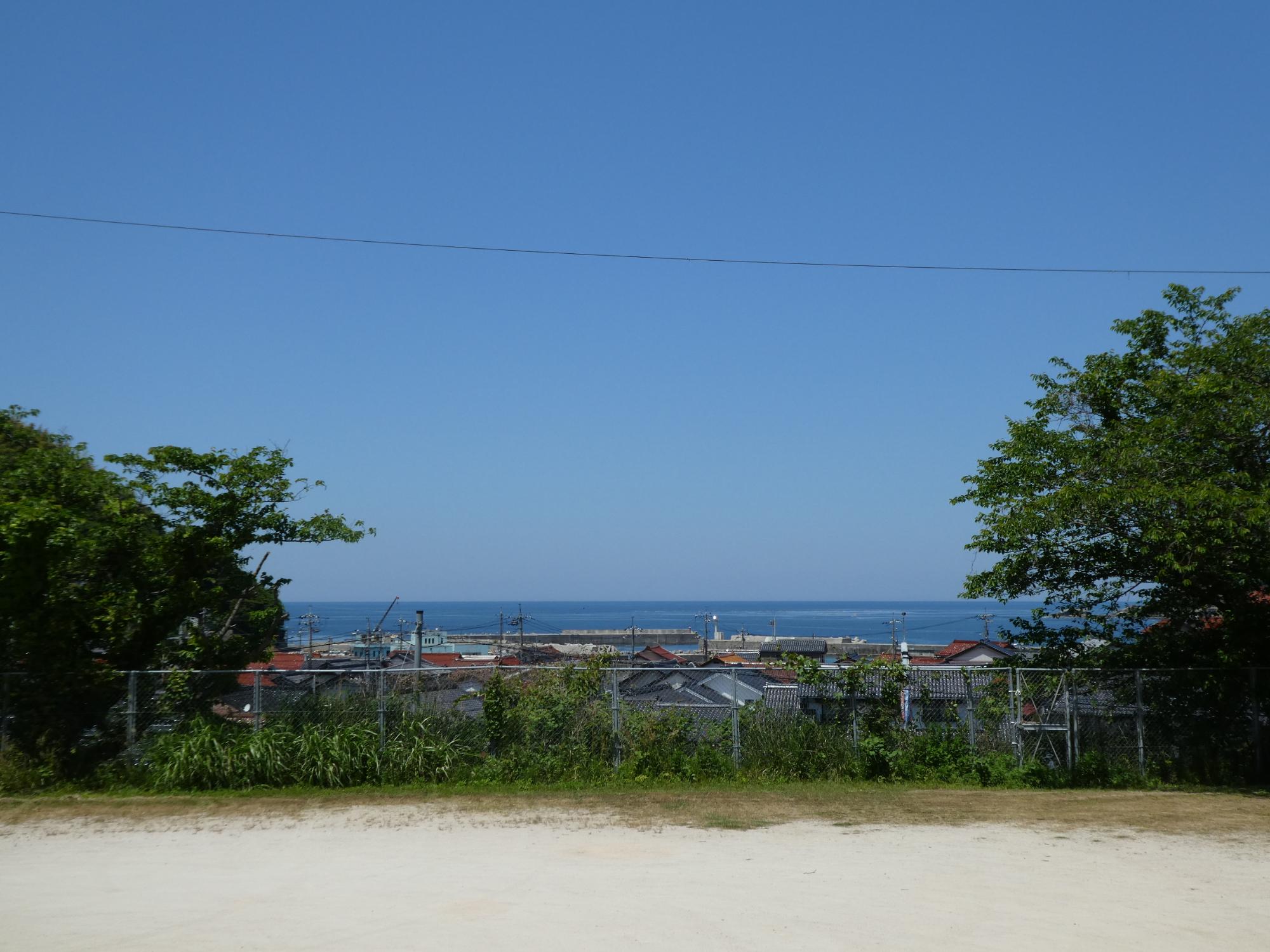 正面奥に青空と水平線があり、左奥に緑の生い茂った山がある。画像中央に御津の港と赤と黒の瓦屋根の町並み。手前の中央にグラウンドとフェンス、左右両脇に緑の葉が茂った木がある。