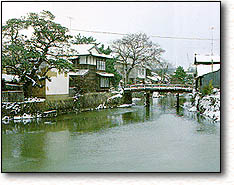 川沿いに樹木と盾門が並び雪が降り積もっている風景写真