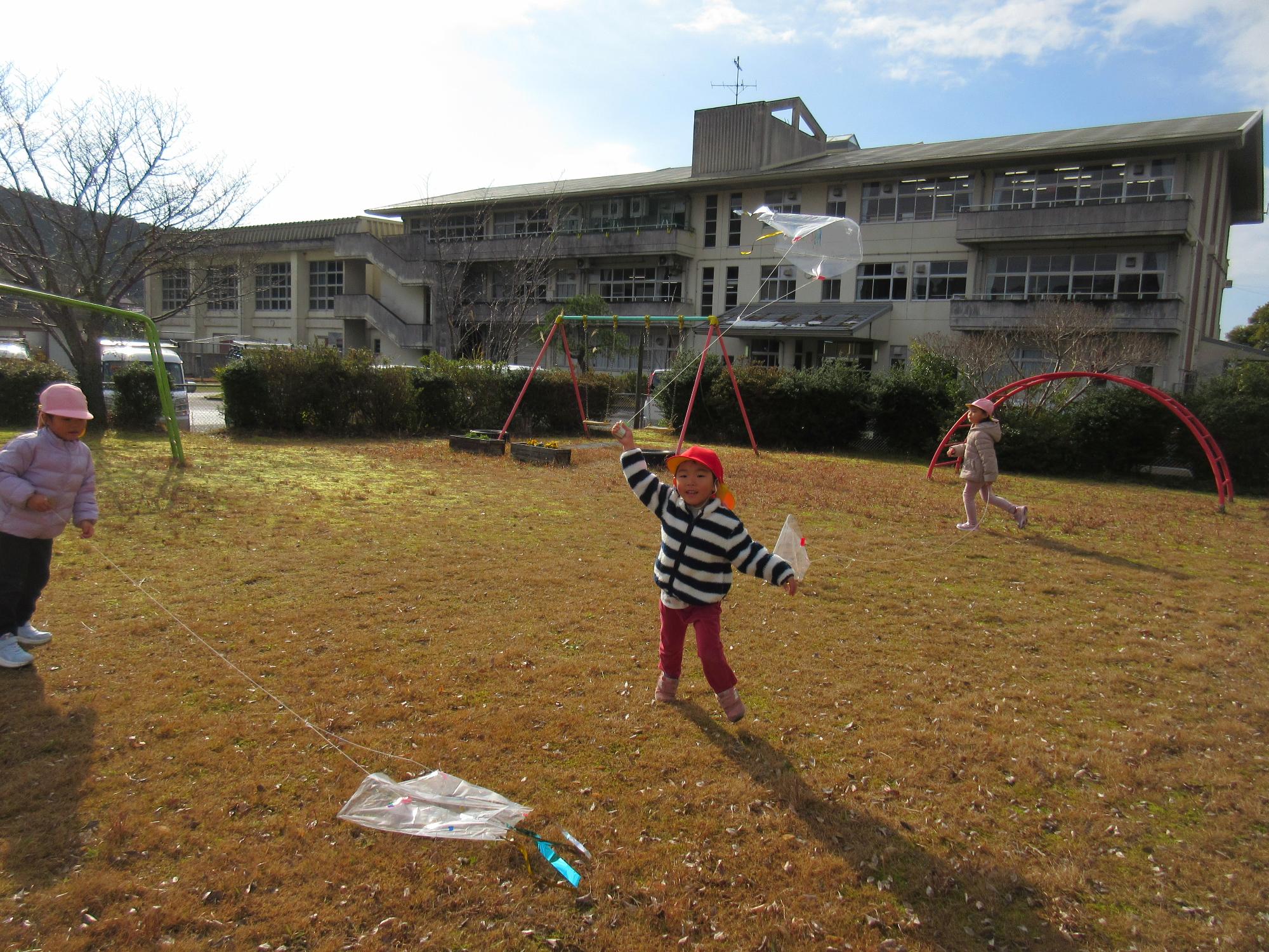 園庭で、こども達が、ビニール袋で作ったたこをあげて遊んでいるところ