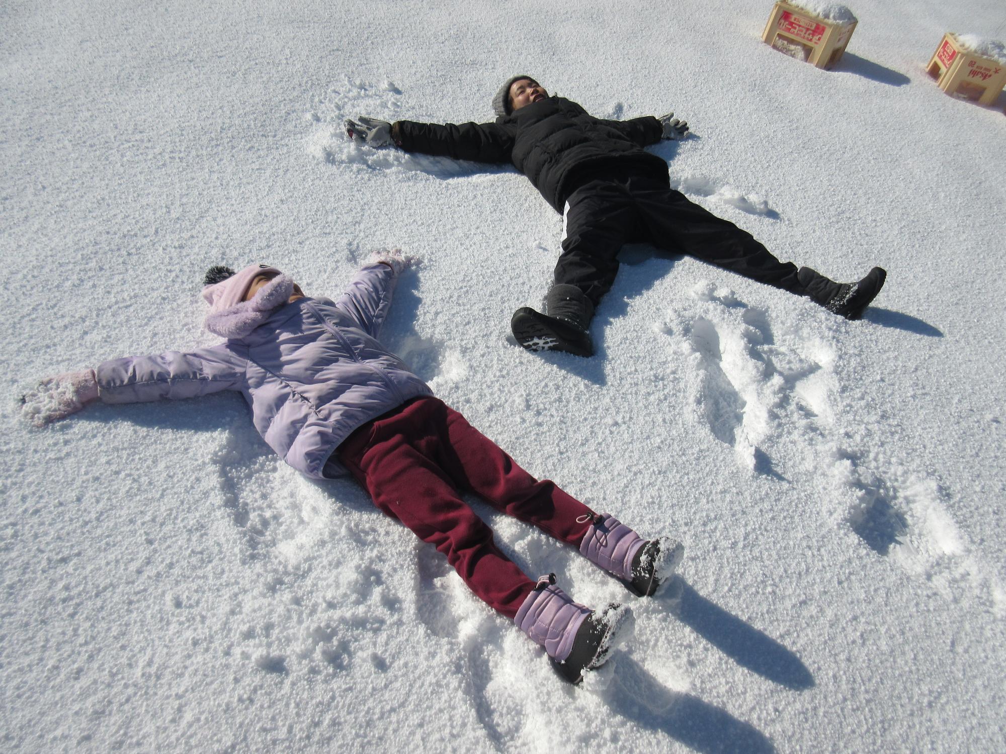 園庭で先生と一人のこどもが、雪の上で大の字に寝転がっているところ
