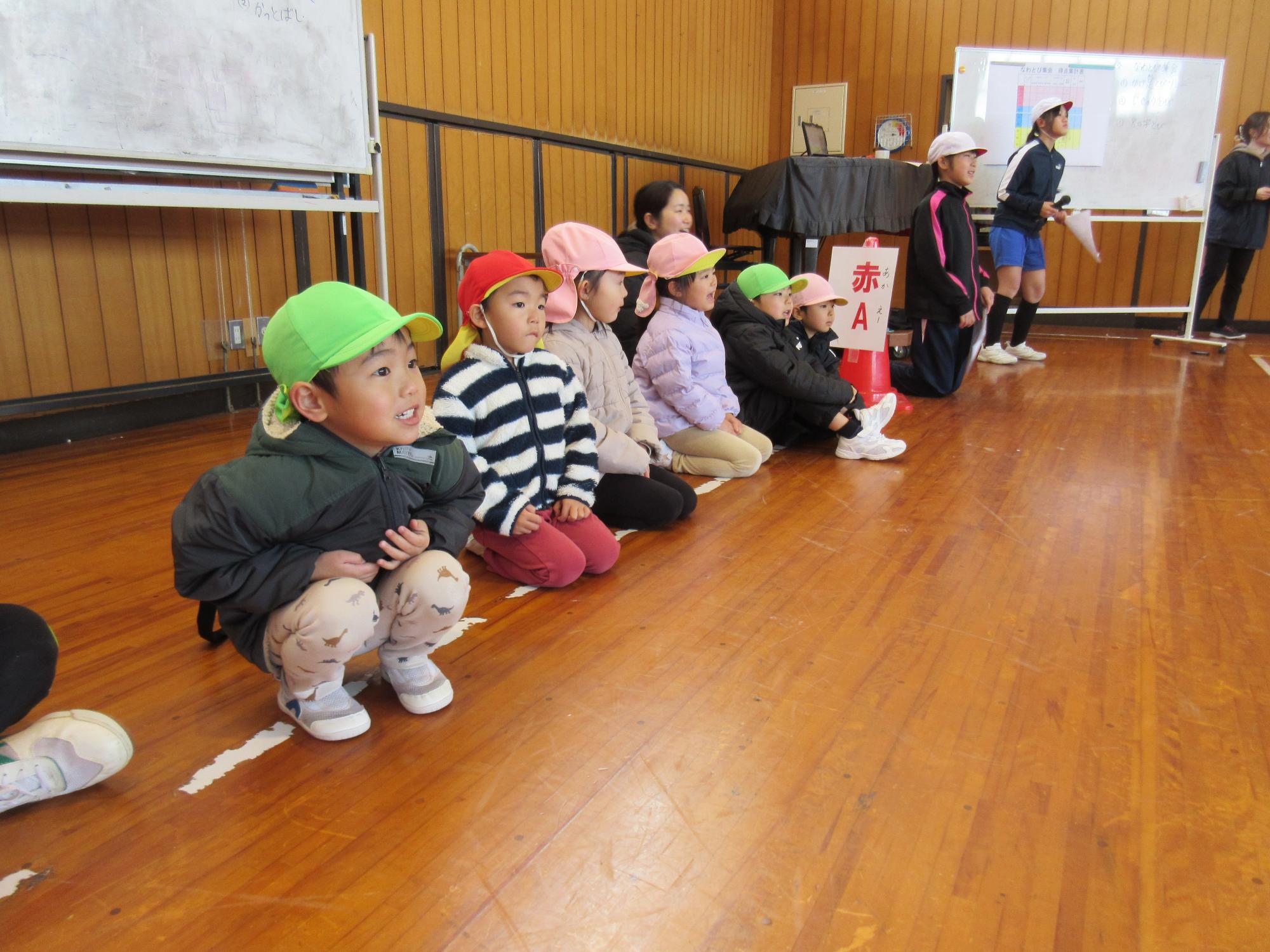 幼稚園のこども達が小学校の体育館で一列に並んで座り、小学生の縄跳びリレーを見ている