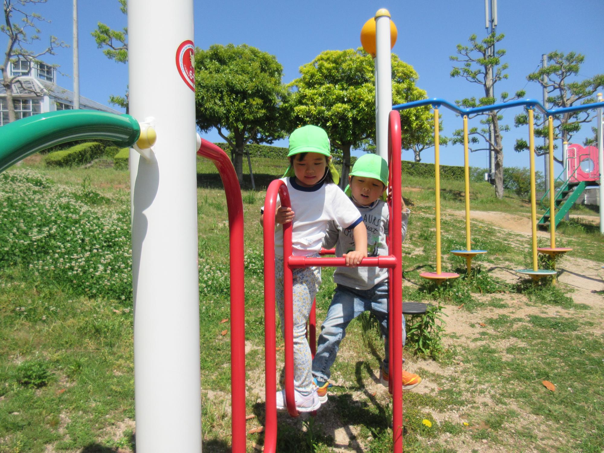 遠足で出かけた公園で、二人のこどもが、棒につかまったり足を開いて渡ったりしながら遊具で遊んでいる。