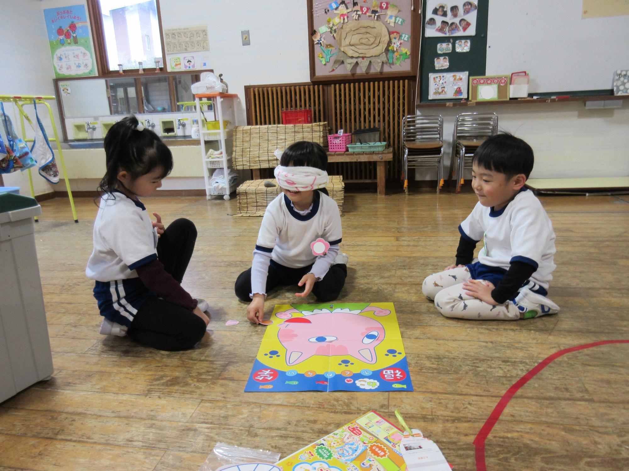 保育室で3人のこども達が、ネコの福笑いをしている。真ん中のこどもがタオルで目隠しをし、他の二人が見ている。
