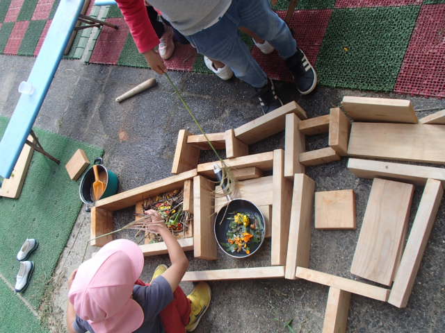 木の板で枠のようなものを作りその中にはっぱを入れたフライパンを置き、おままごとをする園児たちの写真