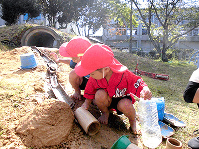 砂場で水を使って遊んでいる赤い帽子をかぶった園児の写真