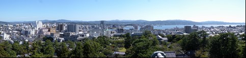 前方に青々とした木々が立ち並び、後方にビルや住宅などが建っている松江城からの眺望写真
