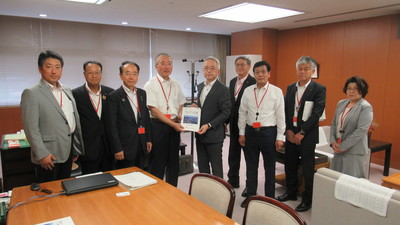 8名の男性と1名の女性が横一列に並び、中央に立つ国土交通省野村正史大臣官房長と同盟会の男性が一緒に要望書を持っている集合写真