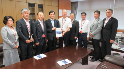 8名の男性と1名の女性が横一列に並び、中央に立つ由木文彦国土交通審議官と同盟会の男性が一緒に要望書を持っている集合写真